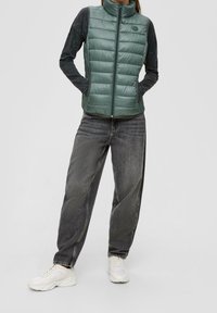 Grön vadderad väst med hög krage och dragkedja, bärs över en mörk långärmad skjorta, i kombination med löst sittande grå jeans och vita sneakers.