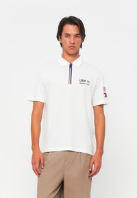 Hvid polo skjorte med lynlås krave, med marineblå og røde detaljer, "USA SAIL GP TEAM" broderi på brystet, og et flag detalje på ærmet.