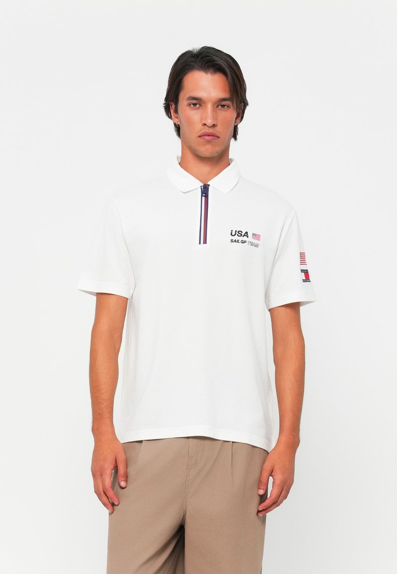 Hvid polo skjorte med lynlås krave, med marineblå og røde detaljer, "USA SAIL GP TEAM" broderi på brystet, og et flag detalje på ærmet.