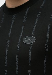 Czarny T-shirt z subtelnym, błyszczącym logo EA7 Emporio Armani oraz powtarzającym się wzorem tekstu w szarości, posiadający okrągły dekolt i gładką fakturę.