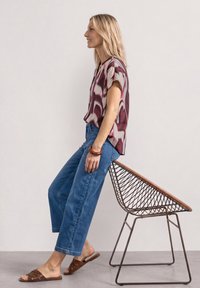 Femme aux cheveux blonds portant un chemisier à motifs, un jean bleu et des sandales marron, assise de profil sur une chaise en fil métallique contre un mur uni.