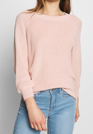 Frau trägt einen hellrosa gestrickten Pullover mit langen Ärmeln und hellblauen Jeans vor einem einfarbigen Hintergrund.
