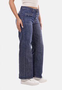 Jeans in denim blu a gamba larga con vita alta, chiusura frontale con bottoni, due tasche laterali e cuciture a contrasto, abbinati a sneakers bianche.