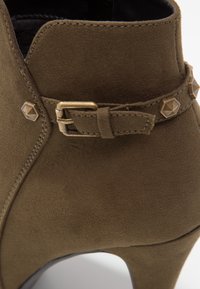 Botte chevillère en suède vert olive avec un talon effilé, dotée d'une sangle avec boucle et d'accents en métal pour plus de détails et de texture.