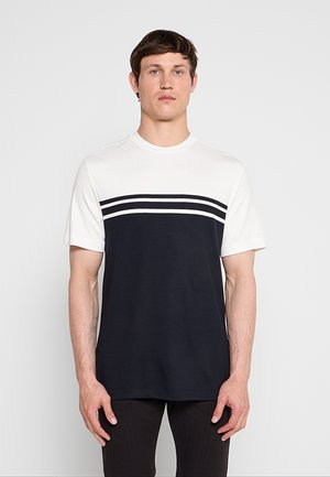 Mand iført en kortærmet hvid og navyblå t-shirt med to hvide vandrette striber henover brystet, står mod en ensfarvet baggrund.