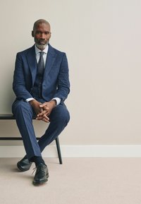 Costum cu carouri bleumarin, cu un design din trei piese, cămașă albă și cravată închisă. Pantofi din piele negri, așezat pe un scaun negru, pe un fundal simplu.