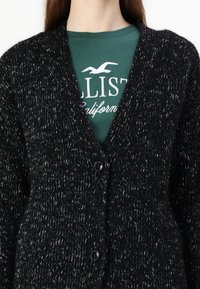 Svart kofta med vita prickar, texturerad stickning, V-ringning och två framknappar. Buren över en grön t-shirt med vit text.