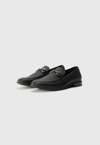 Sorte loafers med teksturert overflate, med en liten stropp og metallkjede aksent. Lav blokk hæl for ekstra komfort.