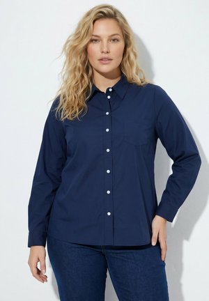 Kvinde med langt, bølget blondt hår iført en mørkeblå skjorte med knapper og mørkeblå jeans, der står mod en hvid baggrund.
