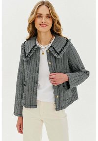 Černobílý kostkovaný kabát s velkým rýškovaným límcem, zlatými knoflíky a dlouhými rukávy. Nosený přes bílé tričko a světlé kalhoty.