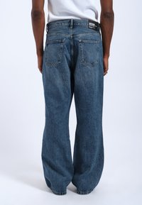 V Loose blue denim-farkut, joissa on haalistunut pinta, viisi taskua, musta brändilogo takana ja näkyvät sauman yksityiskohdat.