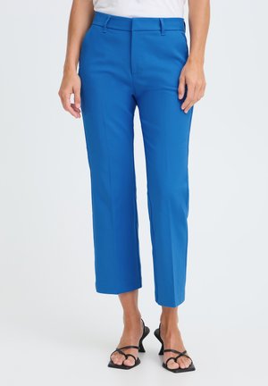 Mujer con pantalones azules brillantes recortados de pierna recta, sandalias negras de tacón y una camiseta blanca sobre un fondo liso.