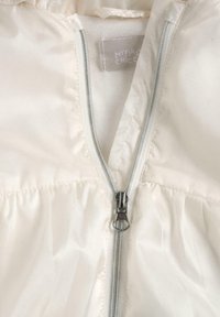 Veste blanche gonflée avec une fermeture éclair frontale, présentant une texture lisse et brillante. La doublure intérieure est douce, avec l'étiquette "MY FIRST CHICCO" visible.