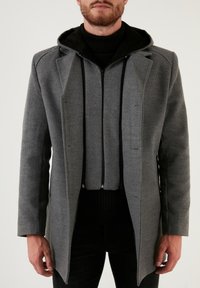 Abrigo gris con solapa estructurada, capucha negra cosida y detalles de cremallera doble. La tela parece suave y texturizada, con un ajuste entallado.