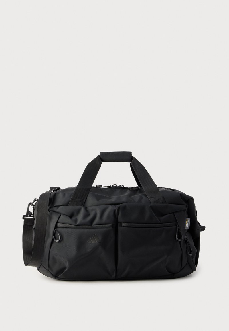 Musta kestävästä nailonista valmistettu duffel-laukku, jossa on kaksi etutaskua vetoketjulla, lyhyet kahvat ja säädettävä olkahihna. Sisältää logon koristeet.