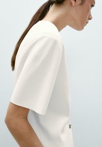 Haut blanc à manches courtes avec un col rond, fabriqué dans un tissu lisse, mettant en valeur des lignes épurées et un détail de bouton unique sur le côté.