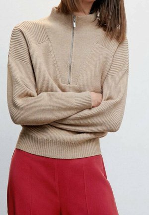 Personne portant un pull beige côtelé avec une fermeture éclair à l'avant et des manches longues, associée à un pantalon rouge taille haute, bras croisés.