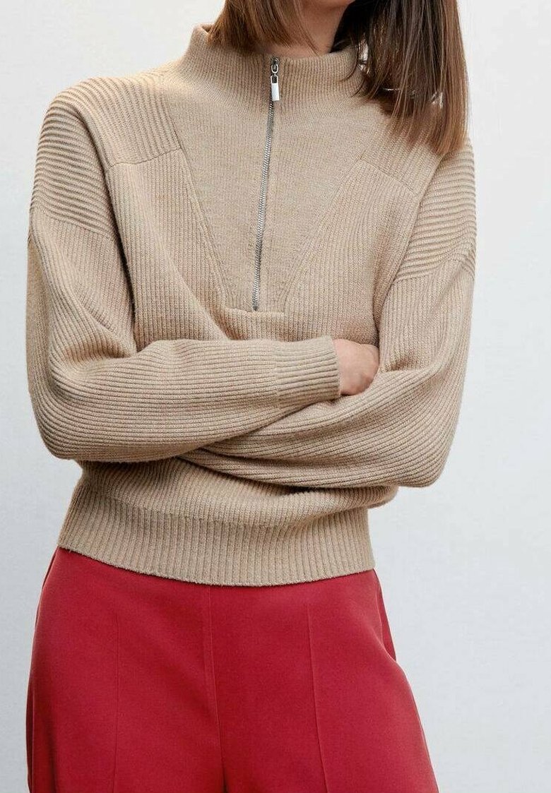 Personne portant un pull beige côtelé avec une fermeture éclair à l'avant et des manches longues, associée à un pantalon rouge taille haute, bras croisés.