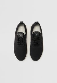 Baskets de sport noires avec une tige en maille, présentant un motif texturé, un système de laçage et une semelle blanche. Étiquette avec le logo sur la languette.