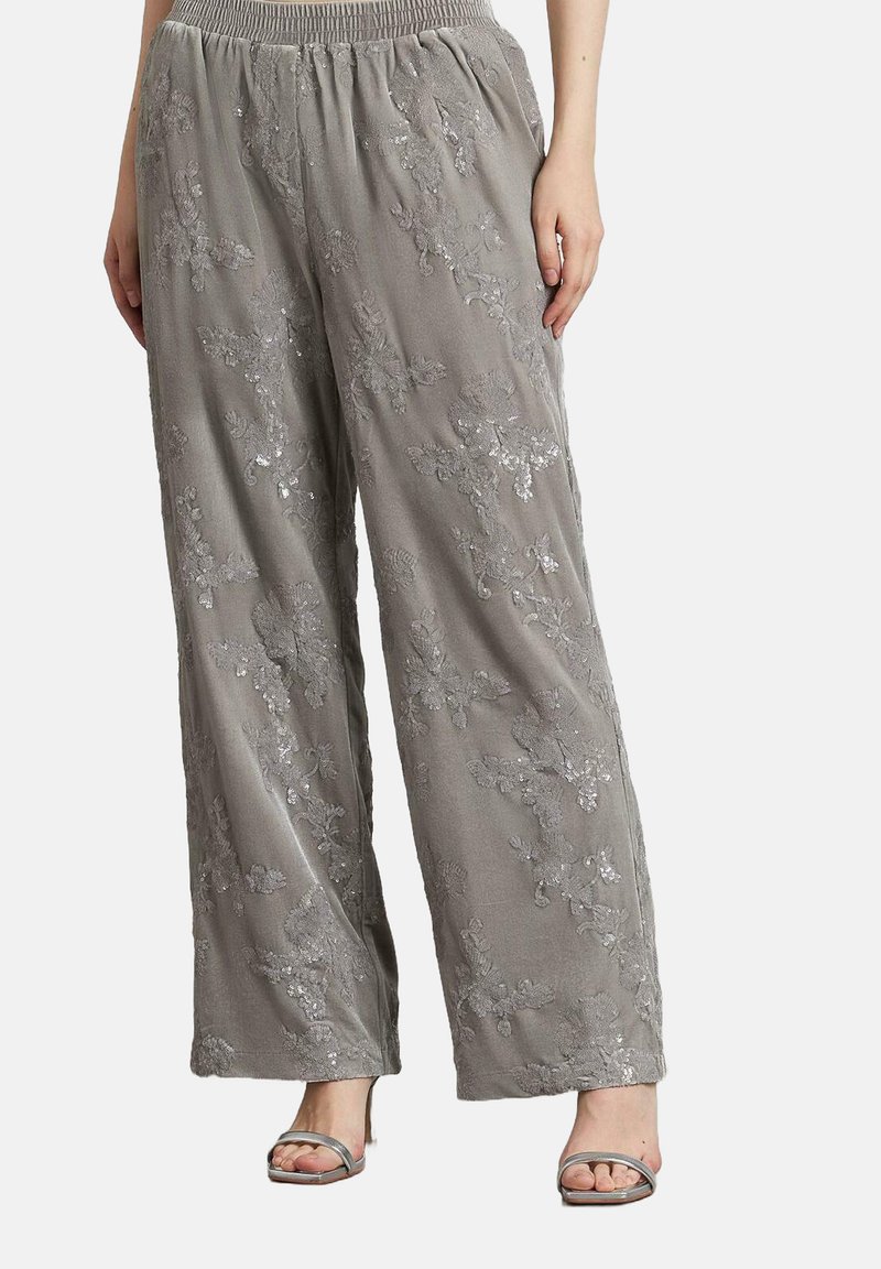 Pantalones anchos grises con bordado de lentejuelas florales, cinturilla elástica, combinados con tacones abiertos plateados sobre un fondo blanco liso.