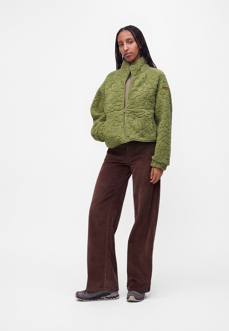 Femme aux longues tresses portant une veste verte texturée zippée, un pantalon marron à jambes larges et des baskets foncées, debout la main dans la poche.