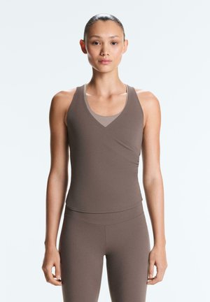 Vrouw staand en recht vooruit kijkend, gekleed in een nauwsluitende, bruine mouwloze top en bijpassende leggings tegen een effen lichte achtergrond.