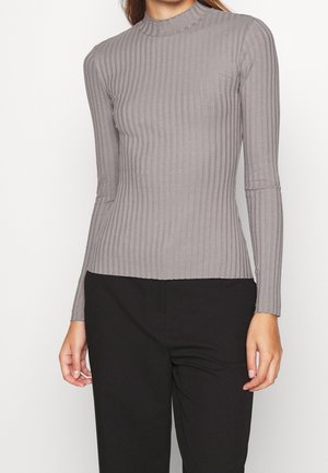 Femme portant un haut gris côtelé ajusté à manches longues avec col montant et pantalon noir, debout devant un fond uni.