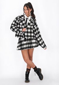 Fekete-fehér kockás, cropped dzseki nagy reverekkel, plisszírozott mini szoknyával párosítva. A modellen fekete fűzős csizmák és egy fehér ing van.