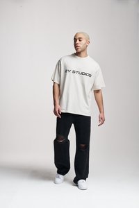 Beige oversized t-skjorte med "2Y STUDIOS" i svart skrift, parret med svarte ripped jeans og hvite sneakers. Enkelt bakgrunn.