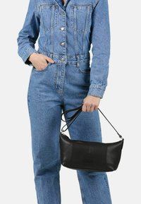Persona in tuta di denim blu che tiene una borsa a tracolla in pelle nera per i manici contro uno sfondo semplice.