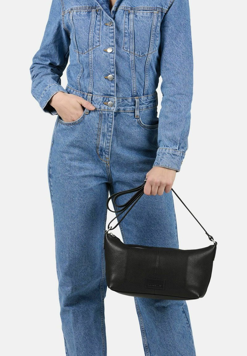 Persona in tuta di denim blu che tiene una borsa a tracolla in pelle nera per i manici contro uno sfondo semplice.