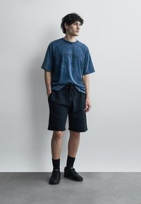 Jeune homme debout contre un mur portant un t-shirt bleu Off-White, un short noir à cordon, des chaussettes noires et des baskets noires, regardant vers la droite.