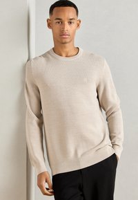 Pull long manches beige clair avec un col rond et des poignets côtelés, présentant un motif de tricot texturé et un logo subtil sur la poitrine.