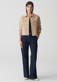 Beige wol cropped jack met voorzakken, knoopsluiting, gedragen over een zwarte top, gecombineerd met donkerblauwe uitlopende jeans en witte schoenen.