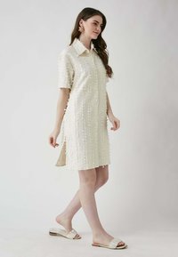 Cremfarbendes Hemdkleid mit kragenförmigem Ausschnitt, kurzen Ärmeln, durchgehender Knopfleiste und seitlichen Schlitzen. Getragen mit hellfarbenen Riemchensandalen.