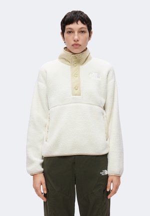 Person iført en cremefarvet fleece-pullover med en beige trykknap-krave og olivengrønne bukser, stående mod en ensartet hvid baggrund.