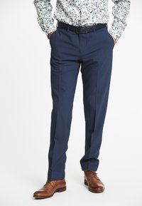Marineblaue maßgeschneiderte Hose mit glatter Textur, seitlichen Taschen und geradem Bein, kombiniert mit braunen Lederschuhen.