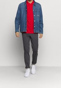 Denim jacket met drukknoopsluiting, rode polo, grijze broek en witte sneakers. De jas heeft een borstzak en een ontspannen pasvorm.