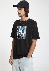 Sort bomulds t-shirt med et stort grafisk design i blå, hvid og sort. Båret sammen med lyseblå denim shorts; modellen står med hænderne i lommerne.
