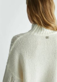 Blondýna v bílém pleteném roláku se malou stř�íbrnou etiketou na horní části zad.