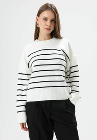 Witte trui met horizontale zwarte strepen, geribbelde zoom en manchetten, losse pasvorm. Gecombineerd met zwarte broek. Soepele textuur.