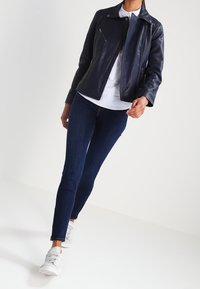 Granatowa skórzana kurtka motocyklowa z detalami na suwak, noszona na białej koszuli, zestawiona z ciemnymi, dopasowanymi jeansami i szarymi trampkami. Gładka faktura.