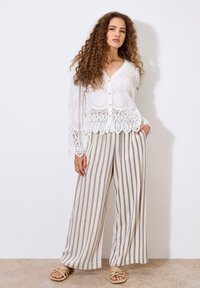 Weiße Spitzenbluse mit floralem Muster und gewelltem Saum, kombiniert mit beige-weißen, gestreiften, weit geschnittenen Hosen. Sandalen mit verdrehten Riemen.
