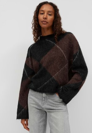 Woman with curly hair wearing a fuzzy black and dark brown checkered sweater and light gray jeans, standing with one hand in pocket.
