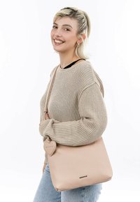 Beige leather crossbody bag with a soft texture, featuring a heart-shaped charm and a minimalist design. Model wears a light sweater and jeans.