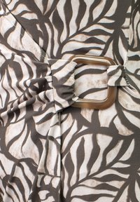 La tela marrón y crema presenta un patrón de hojas, con un detalle fruncido asegurado por una hebilla cuadrada de plástico transparente.