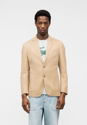 Egy férfi bézs blézert visel fehér grafikás póló fölött és világoskék farmert, egy egyszerű világos háttér előtt áll.