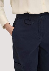 Vrouw in een donkerblauwe, op maat gemaakte broek met zijzakken en een lichtbeige shirt met lange mouwen, hand rustend in de zak.