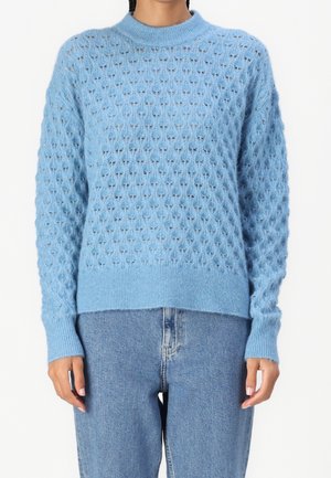 Personne portant un pull tricoté bleu clair avec un motif structuré associé à un jean en denim bleu moyen sur un fond uni.