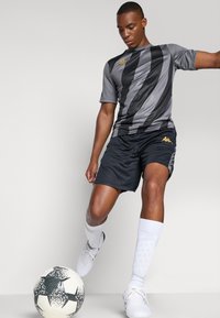 Grå og svart stripet treningsskjorte kombinert med svarte shortser. Modellen har på seg hvite sokker og sko, med en svart-hvit fotball i nærheten.